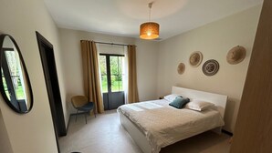 A bedroom with a bed, a chair, a mirror, a window with curtains, and a hanging light fixture. - Maison Proche lac Hourtin et Océan (Gaillan-en-Médoc)