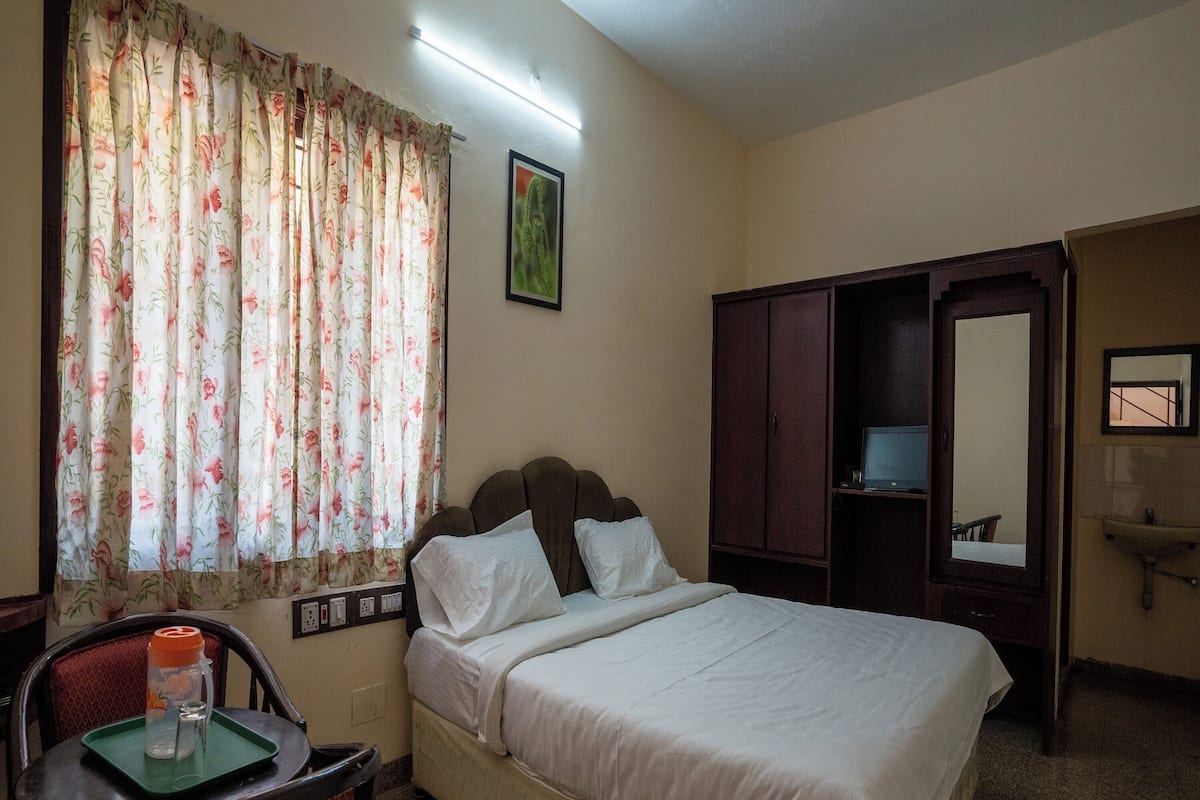 A hotel room with a bed, a chair, a table with a water bottle, a wardrobe, a mirror, and a window with floral curtains.
