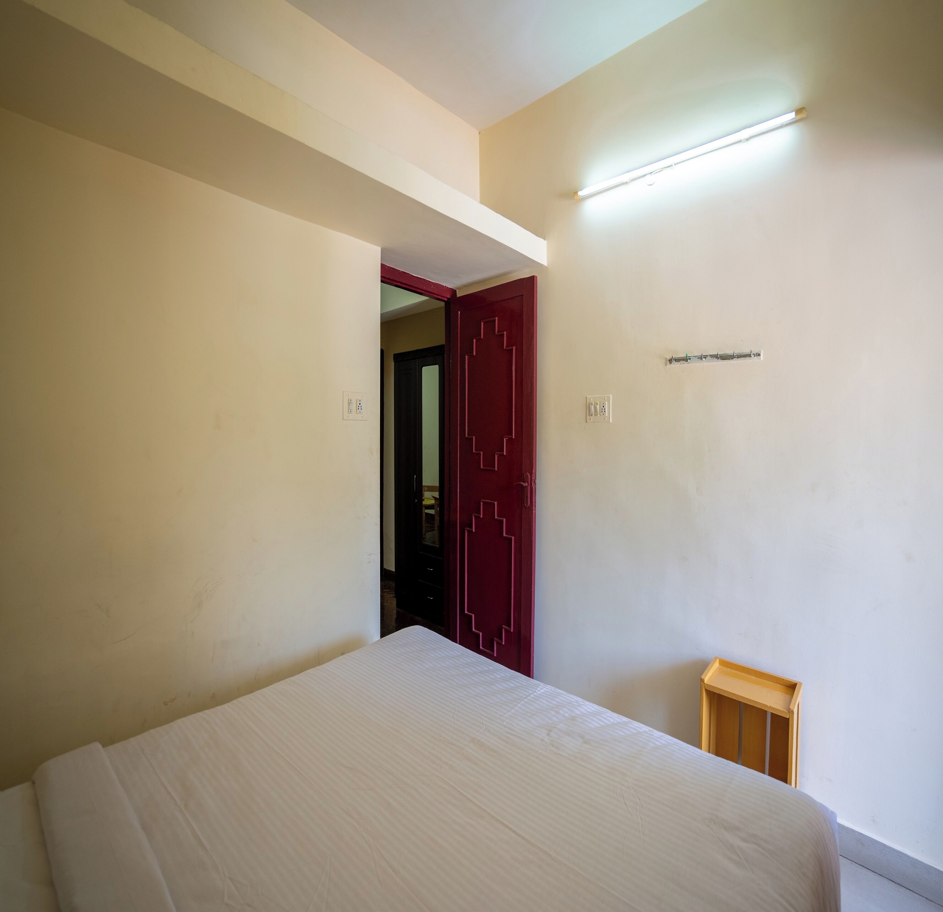 Quarto com uma cama, uma porta vermelha e um banquinho de madeira.
