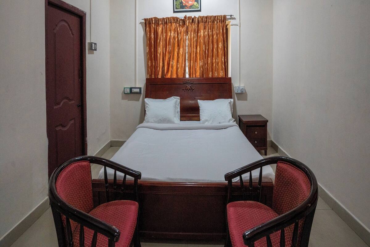 A bedroom with a wooden bed, two red upholstered chairs, a wooden nightstand, and a painting of a flower on the wall.