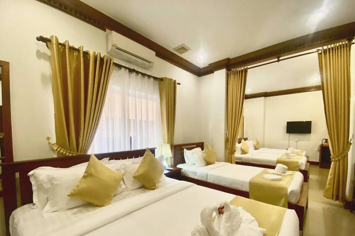 A hotel room with three single beds, each with white linens and gold accents, a flat-screen TV mounted on the wall, and an air conditioning unit above the beds.