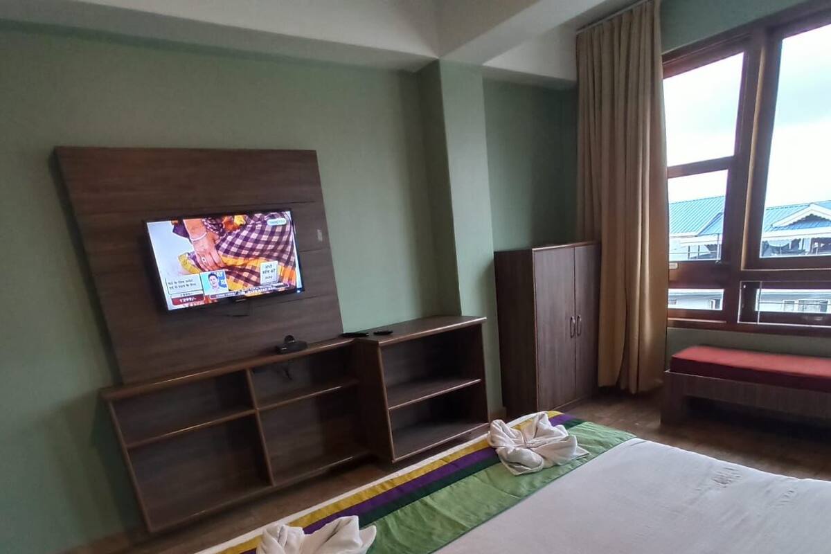 A hotel room with a bed, a TV mounted on the wall, a wooden wardrobe, and a window with curtains.