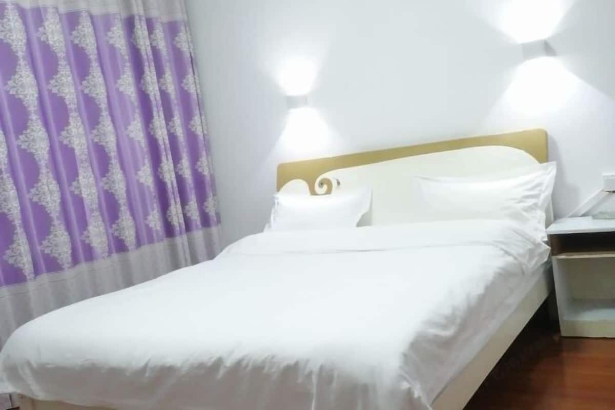 A neatly made bed with white linens and a headboard, a nightstand with a lamp, and a purple and white patterned curtain.
