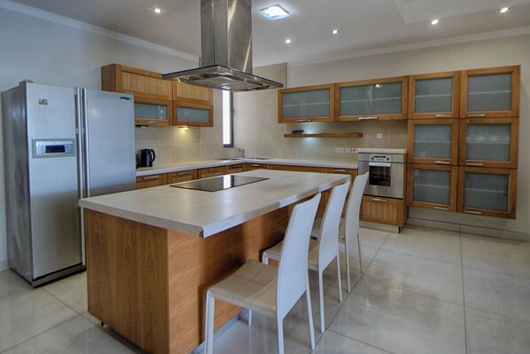 A modern kitchen with a central island, wooden cabinets, stainless steel appliances, and white chairs.
