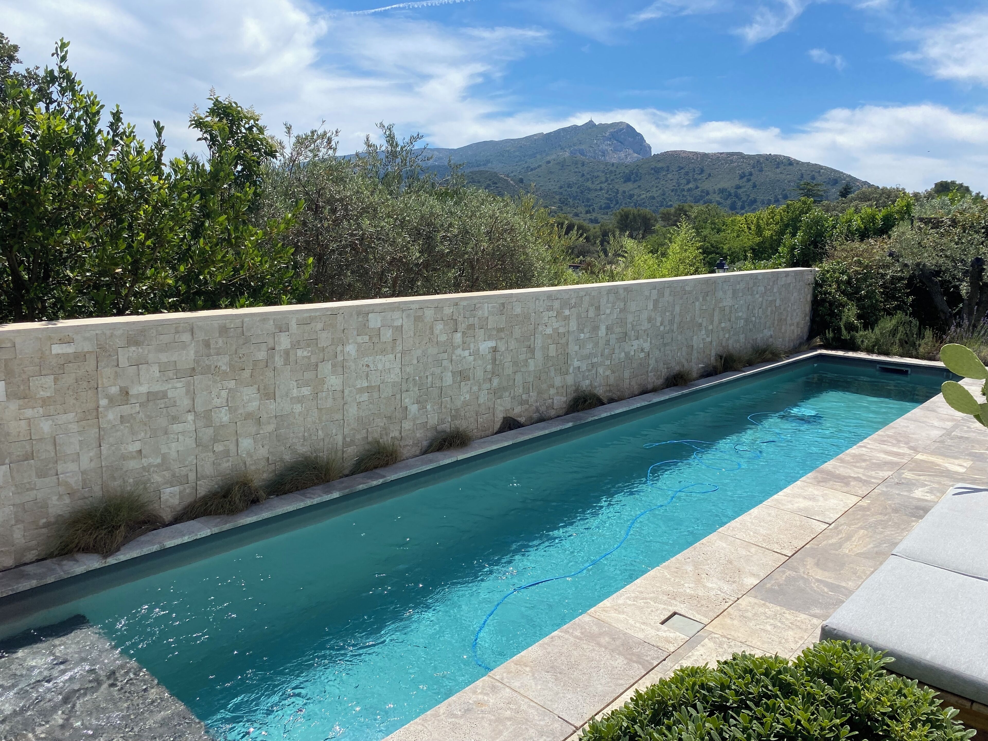 Una piscina rectangular con una pared de piedra en un lateral, rodeada de vegetación y un paisaje montañoso al fondo.