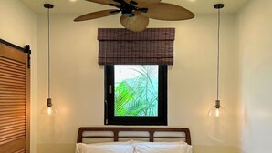 A neatly made bed with a quilted cover, a ceiling fan, and a window with a view of greenery.