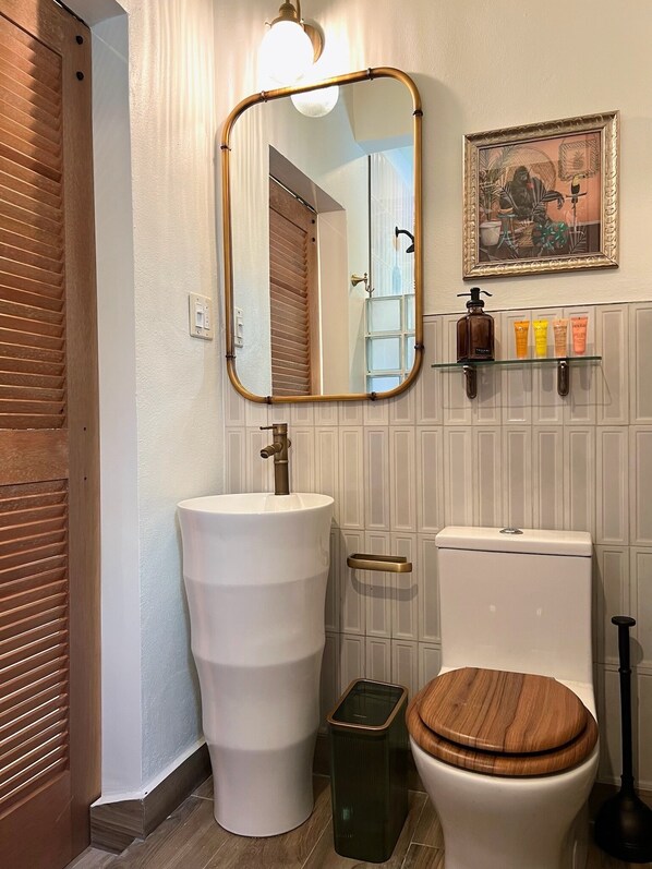 Un baño con un lavamanos grande y blanco, un inodoro con asiento de madera, un espejo con marco dorado y un estante con artículos de tocador.