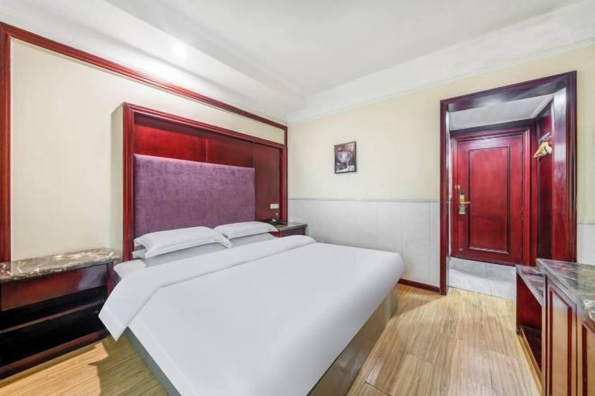 A hotel room with a large bed, wooden headboard, a small bedside table, a nightstand, a red door, and a framed picture on the wall.