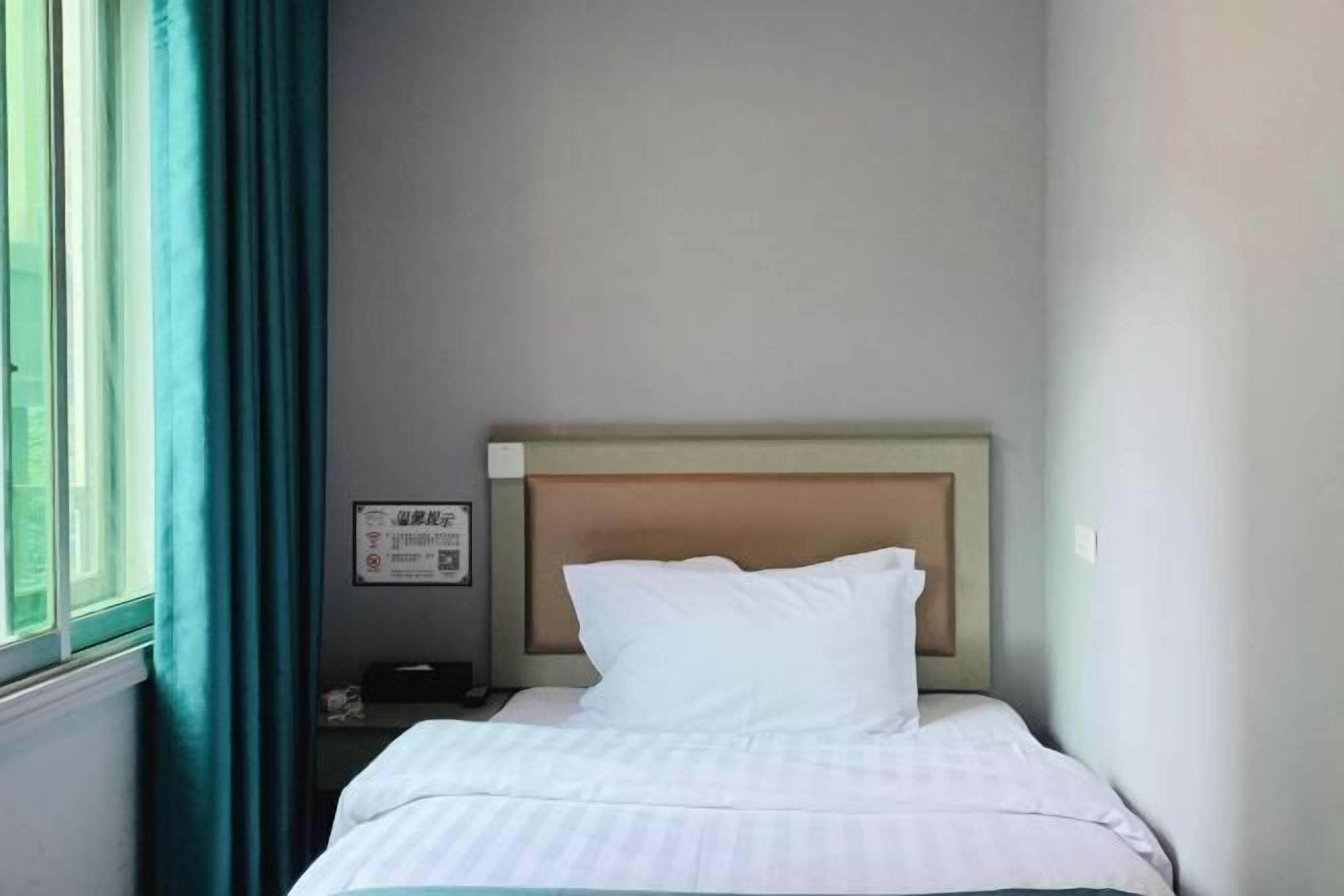 A small, neatly made bed with a wooden headboard in a room with a window and a wall-mounted sign.