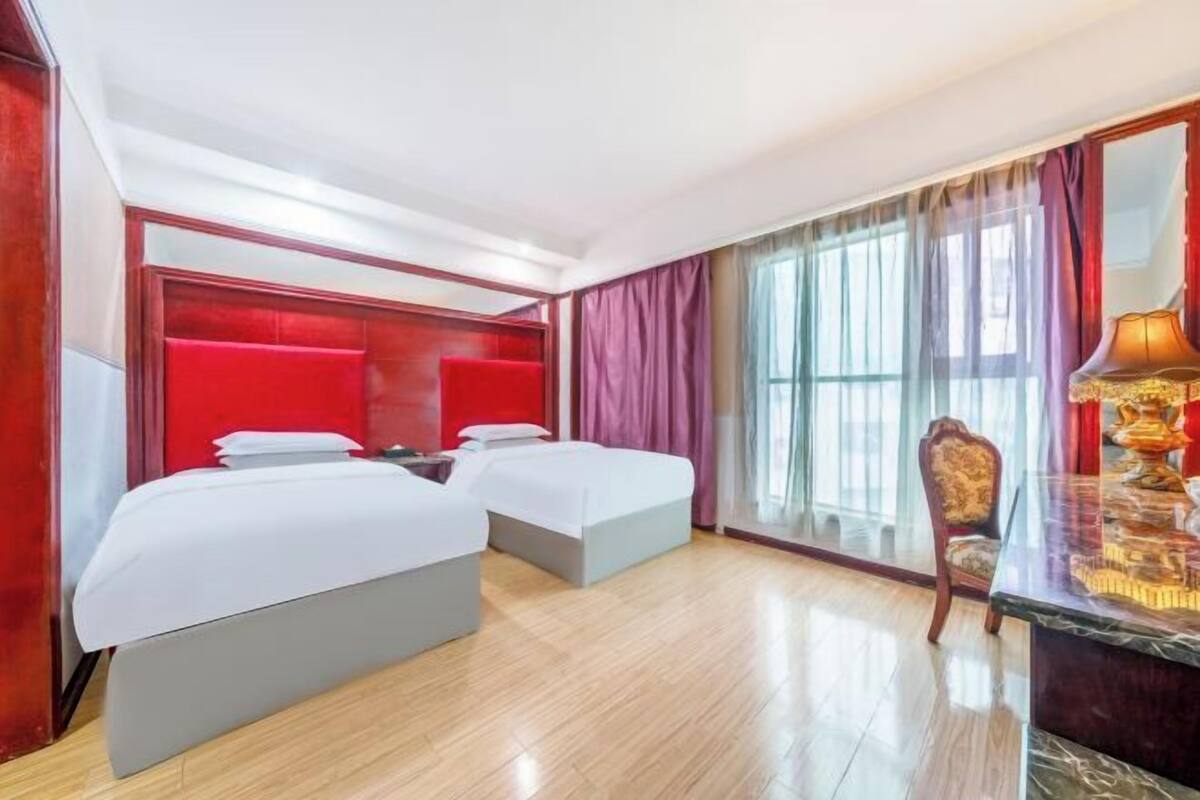 A hotel room with two beds, a wooden desk, and a large window with curtains.