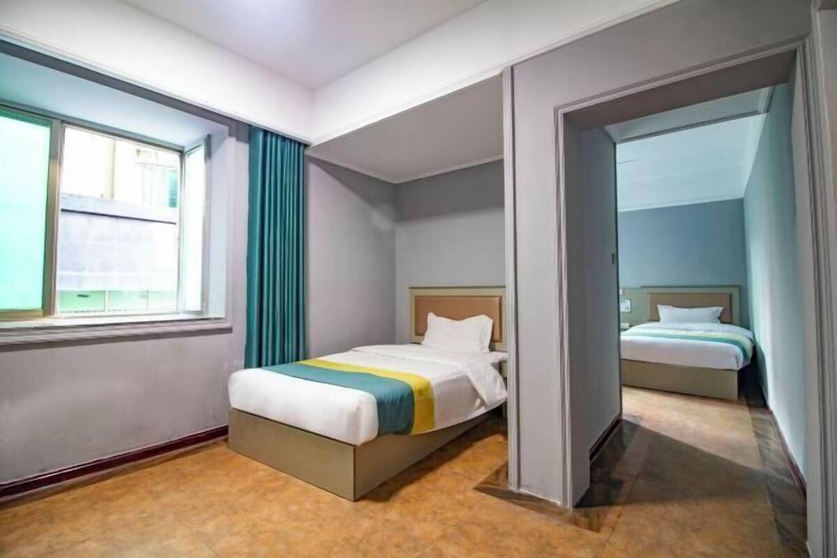 A hotel room with a single bed, a window with curtains, and a view of another room.