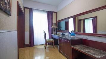 A hotel room with a wooden floor, a desk with a lamp, a chair, a television, and a bench.