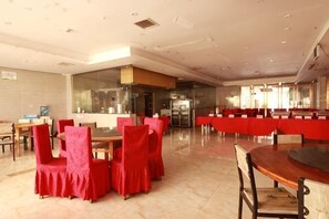 Un grand espace dédié à la salle à manger, avec plusieurs tables et chaises, une cuisine à l’arrière-plan et une grande fenêtre qui laisse entrer la lumière naturelle.