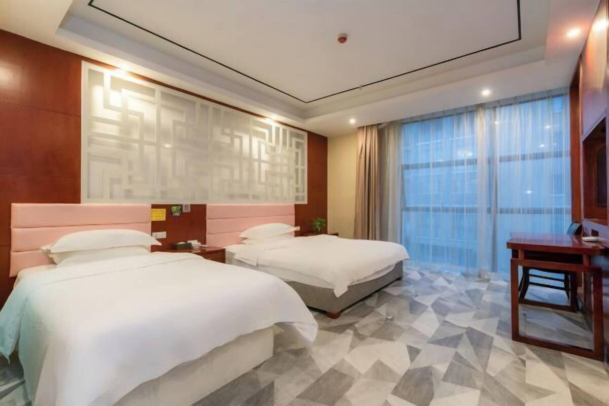 Ett hotellrum med två sängar, ett stort fönster med gardiner, ett golv med ett geometriskt mönster och en sänggavel i trä.