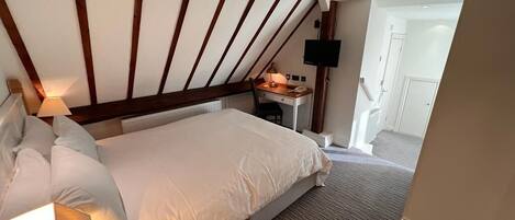 A hotel room with a large bed, a desk with a chair, a flat-screen TV mounted on the wall, and a door leading to a hallway.