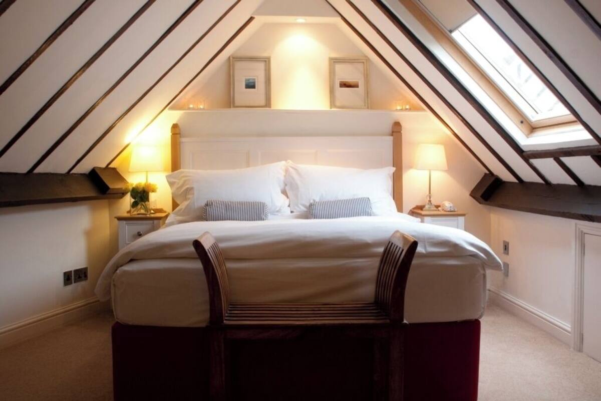 Un dormitorio con techo inclinado, una cama con ropa de cama blanca, dos lámparas de noche y cuadros enmarcados sobre la cama.