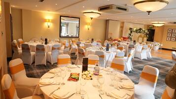 Un salón de banquetes con mesas redondas dispuestas para comer, sillas blancas con cintas anaranjadas y lámparas fijadas a la pared.