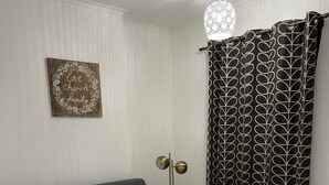 A neatly made bed with white linens, a bedside table with a lamp, a wall decoration, and a patterned curtain. - F2S Serviced 2Bed House Wi-fi, Netflix, Parking, Train station nearby. Holiday! (Derby)