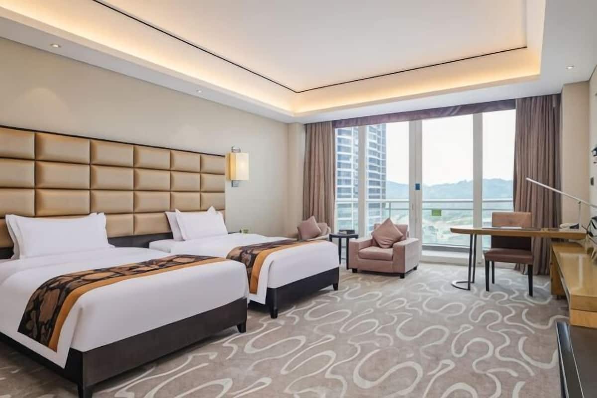 A hotel room with two beds, a desk, a chair, a large window, and a view of the city.