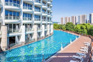 Piscina junto a un edificio moderno con balcones, rodeada de una zona ajardinada con sillones de descanso.