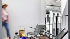 Een balkon met twee grijze lounge fauteuils, een rieten mand, een glas rode wijn en een boek.
