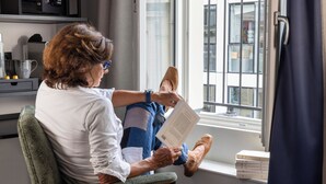 Iemand zit bij een raam met een boek, een stoel en een stapel boeken op de vensterbank.