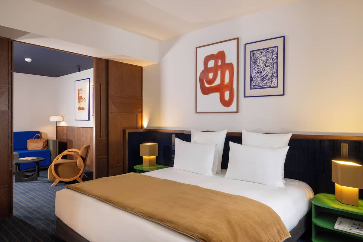 A hotel room with a large bed, two bedside tables with lamps, a wooden chair, a small table with a chair, and artwork on the walls.