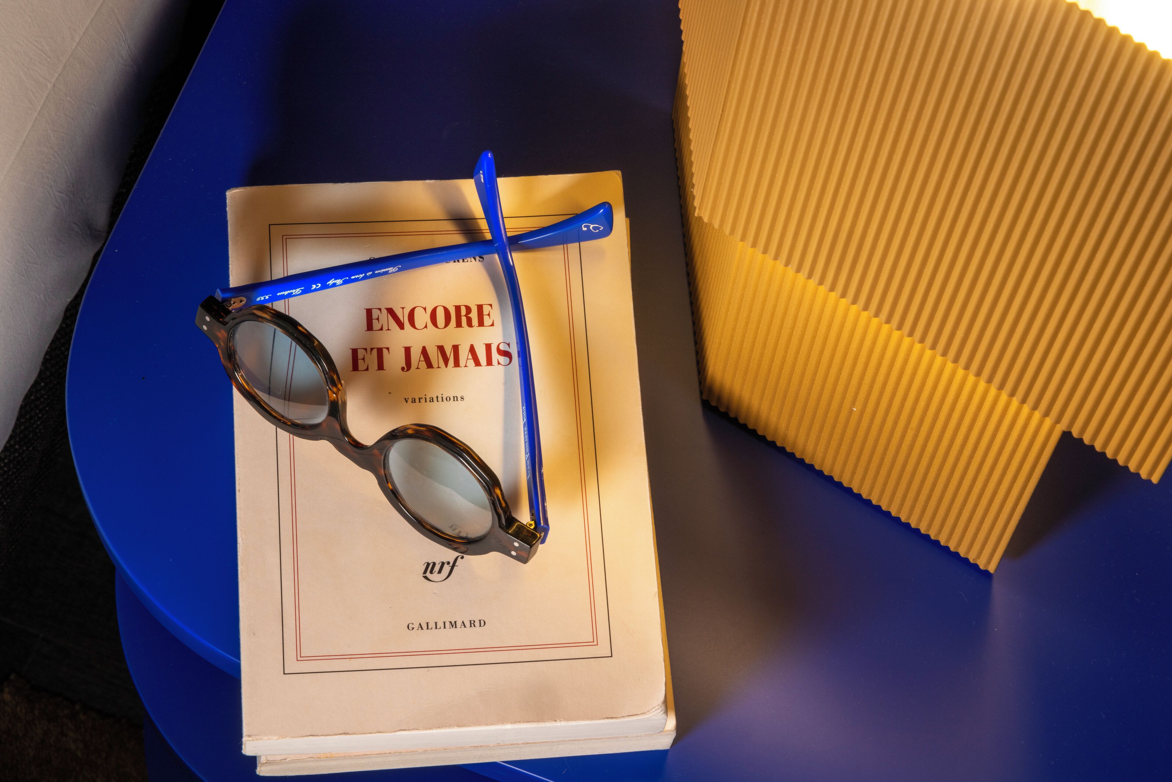Un livre intitulé « Encore et jamais », sur lequel repose une paire de lunettes, est posé sur une surface bleue, à côté d’un objet jaune.