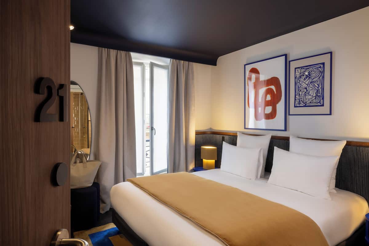 A hotel room with a large bed, a bedside table with a lamp, a bathroom with a mirror, and a window with curtains.