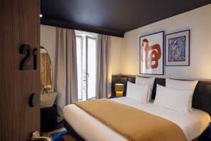 A hotel room with a large bed, a bedside table with a lamp, a bathroom with a mirror, and a window with curtains. - Hôtel Gabbie (Paris)
