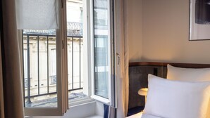 Een hotelkamer met een groot raam, een bed met witte lakens en een nachtkastje met een lamp.
