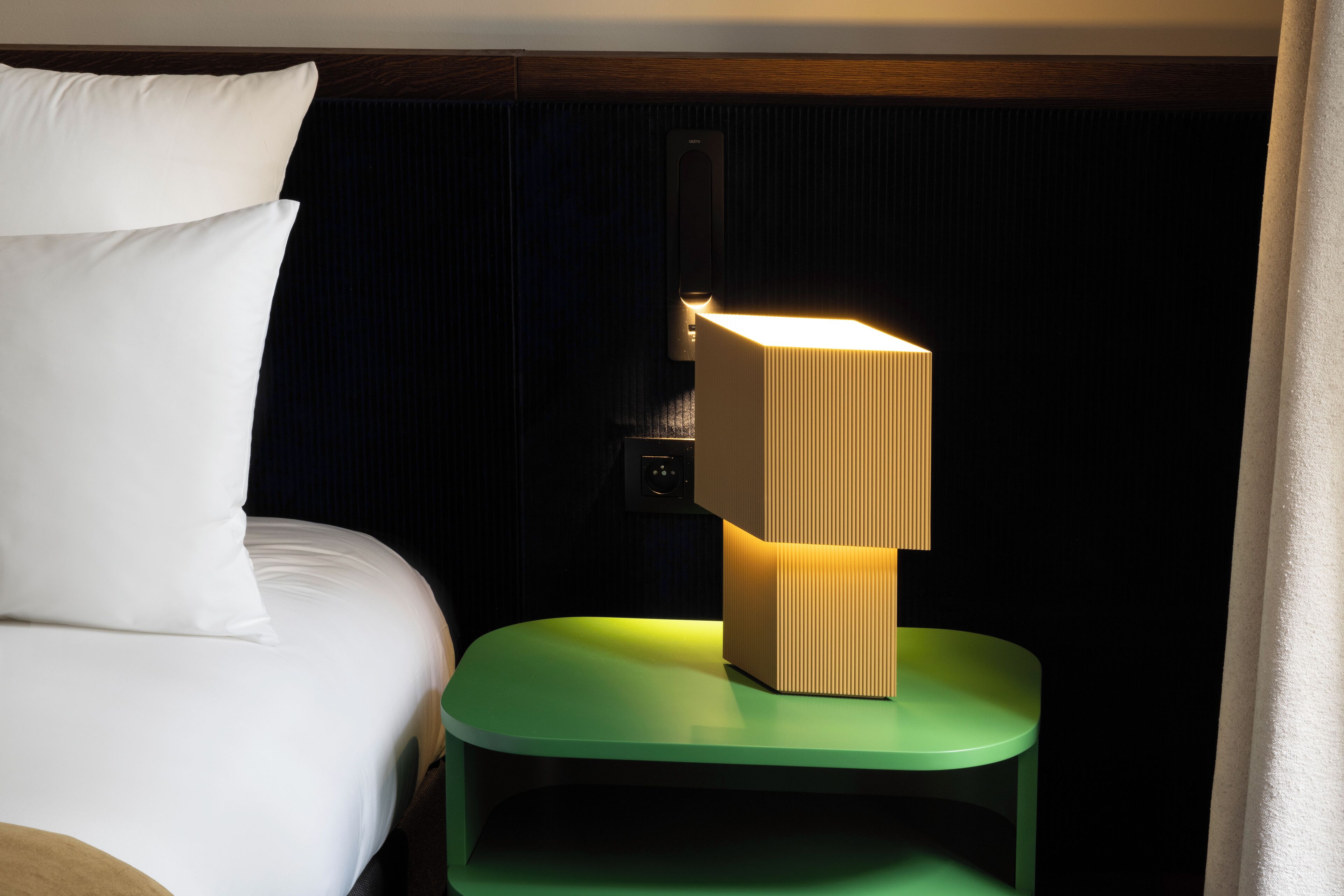 Un lit bien fait, avec une literie blanche, une table de chevet verte sur laquelle se trouve une lampe, et un mur foncé avec un interrupteur.
