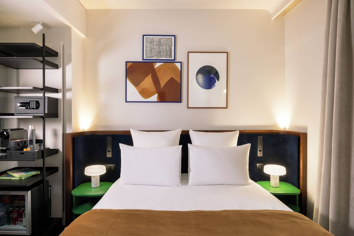 A hotel room with a bed, two bedside tables, a desk with a computer, a mini-fridge, and artwork on the wall.