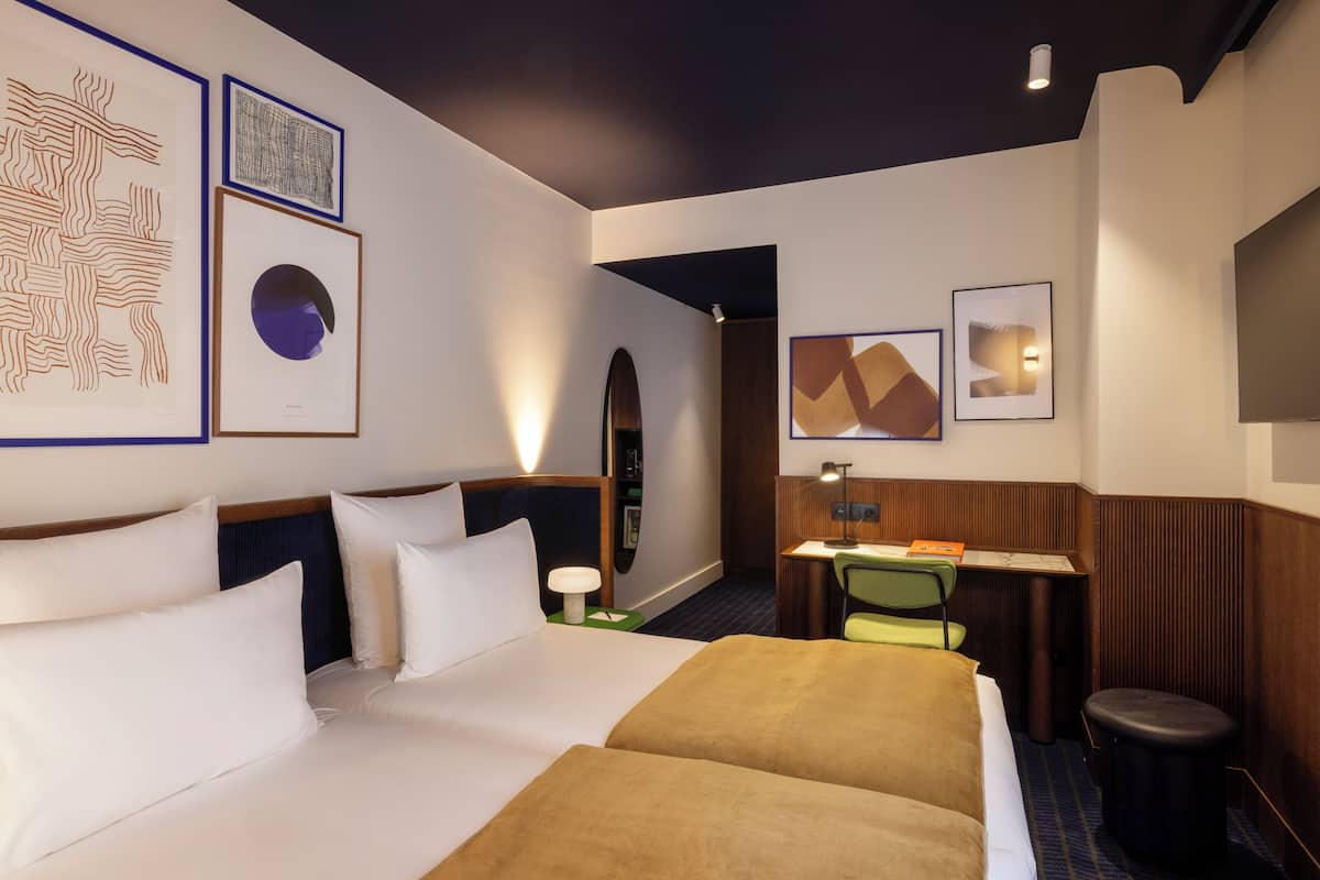 A hotel room with a large bed, a desk, and a chair. There are framed artworks on the walls and a small stool near the desk.