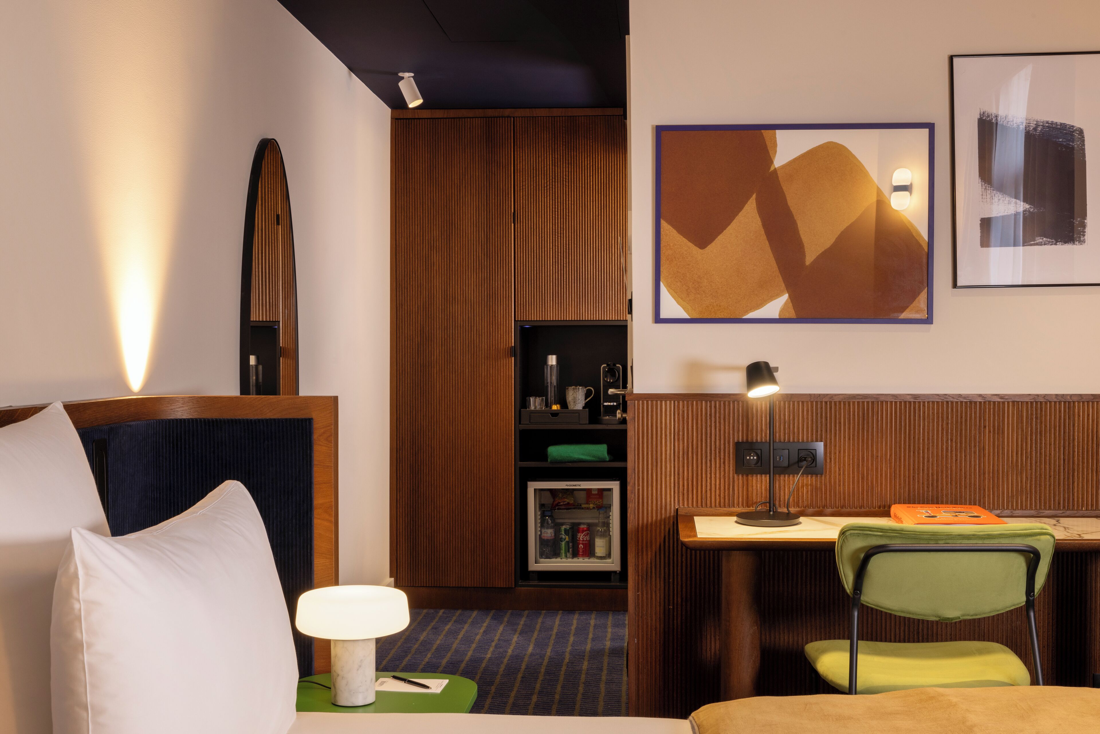 A hotel room with a bed, a desk, a chair, a lamp, a refrigerator, and a wall-mounted shelf.