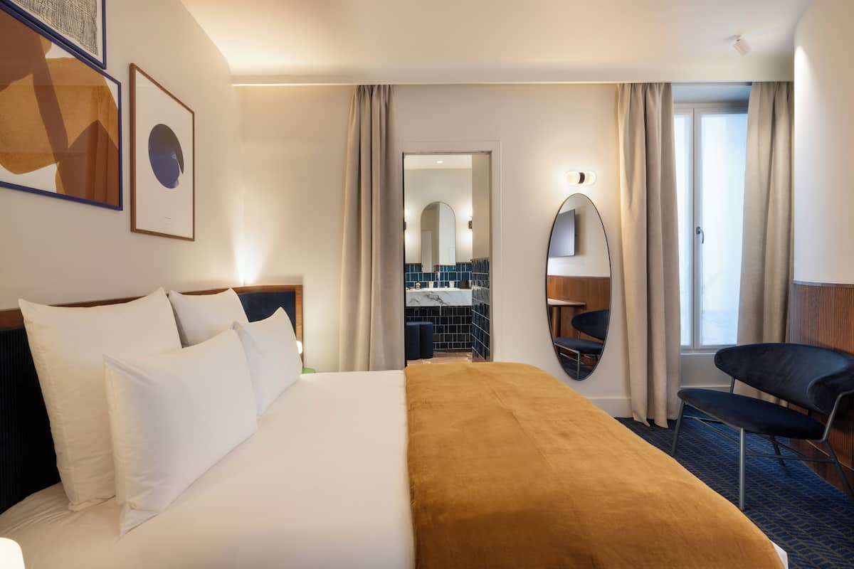 A hotel room with a large bed, a desk, a chair, a mirror, and a bathroom visible through an open door.