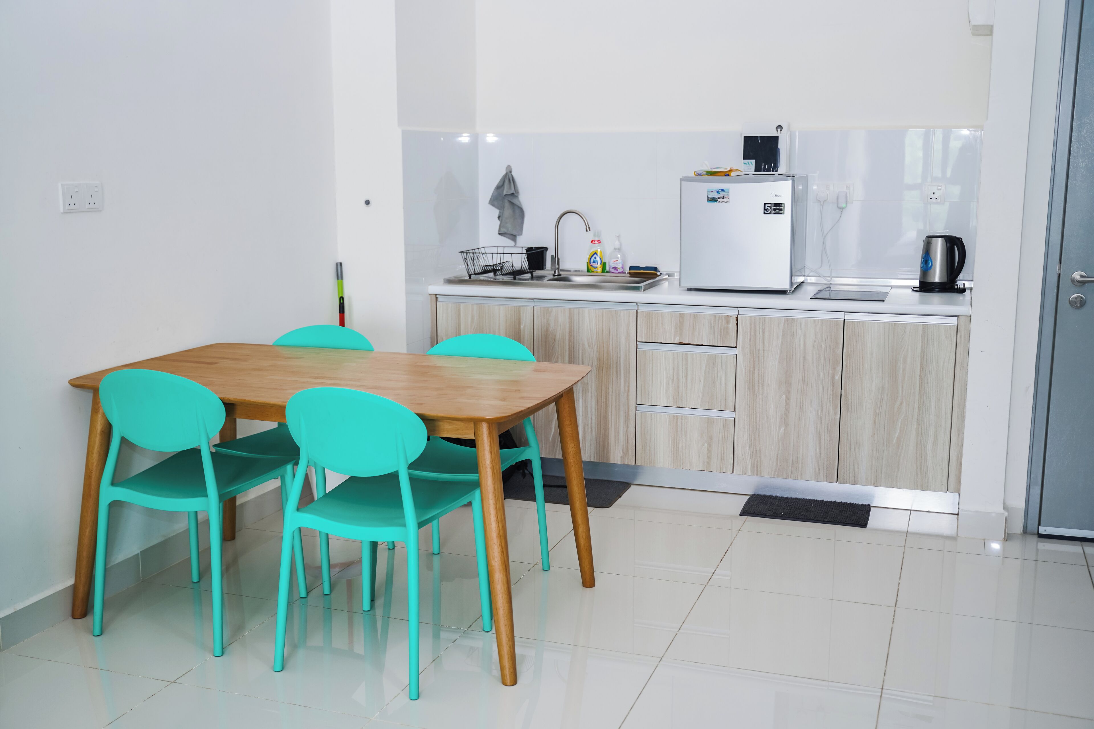 Een moderne keuken met een houten tafel en turquoise stoelen, een witte koelkast en een waterkoker op het aanrecht.