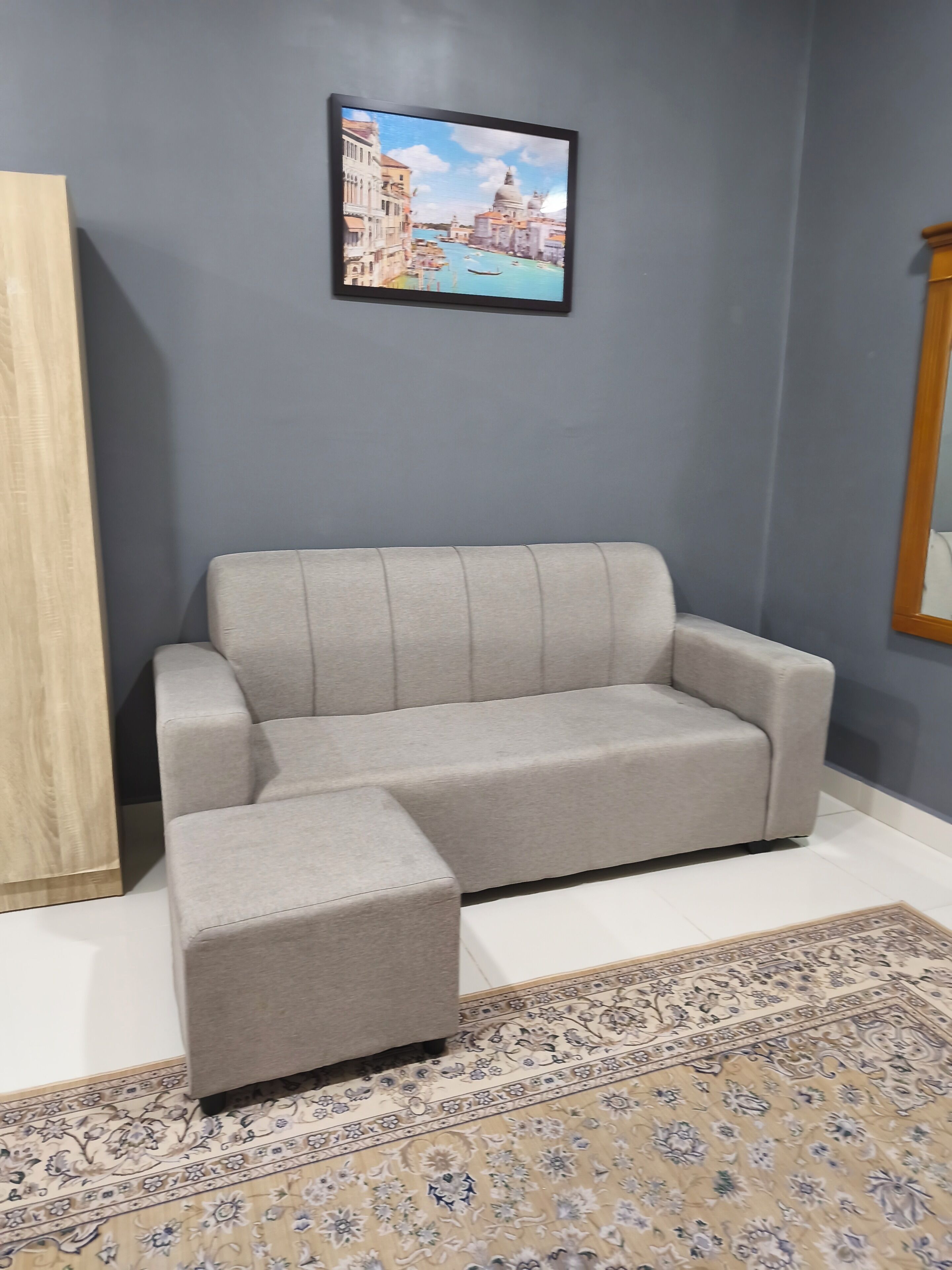 A grey sofa and ottoman in a room with a patterned rug, a framed picture on the wall, and a wooden sideboard.