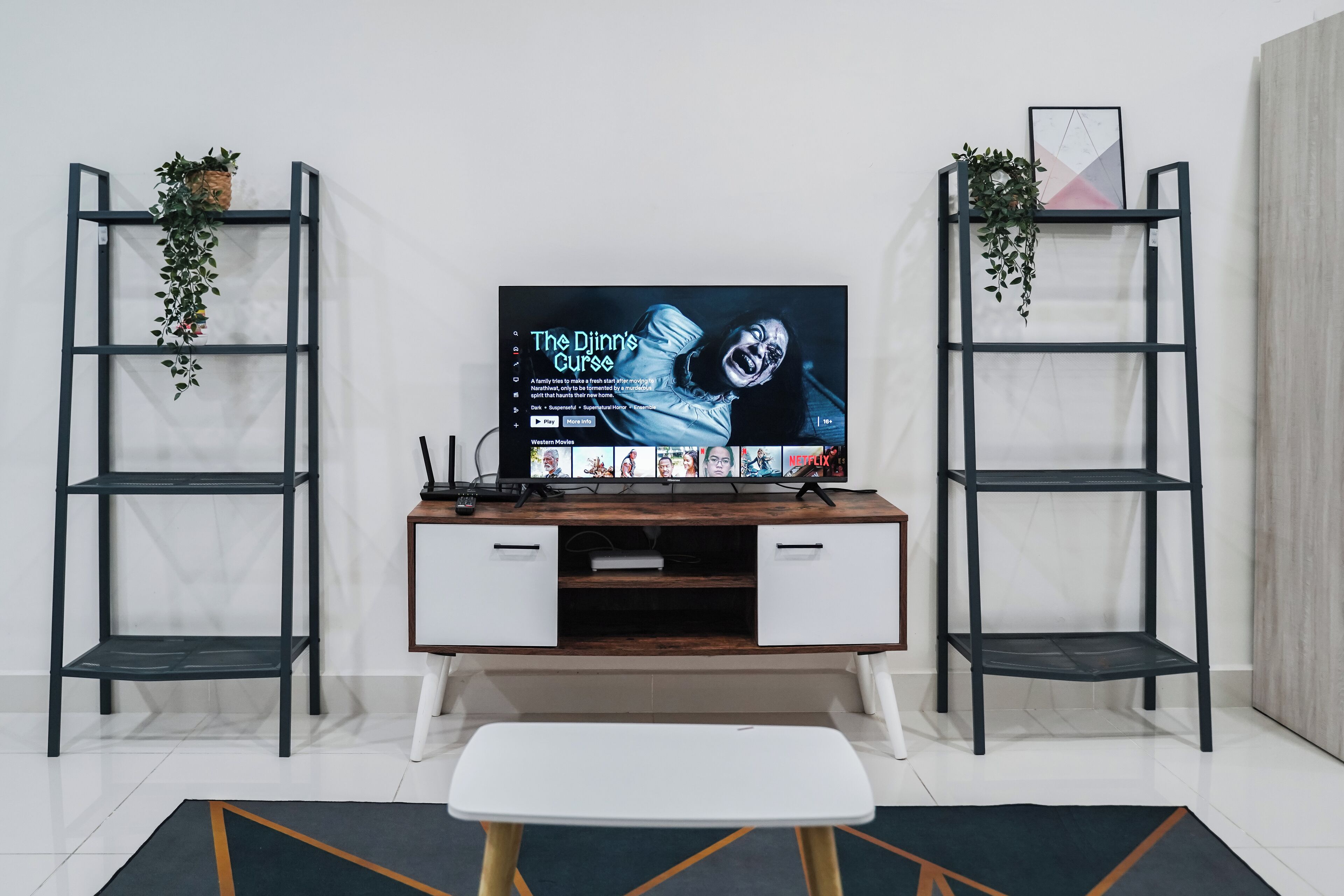 Una sala de estar con un televisor sobre un mueble de madera, dos estantes con plantas y una mesita.