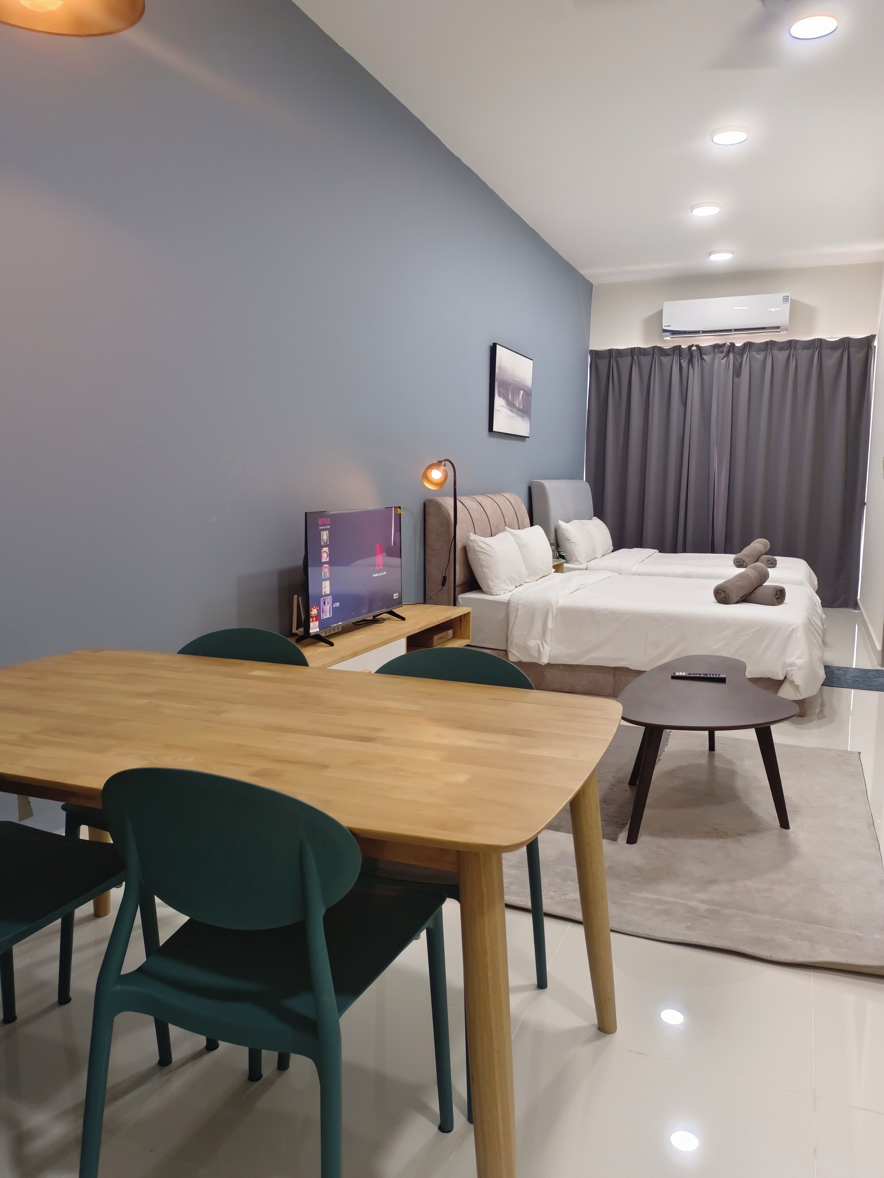 A modern hotel room with a wooden dining table, teal chairs, a bed with white linens, a grey curtain, and a mounted air conditioner.