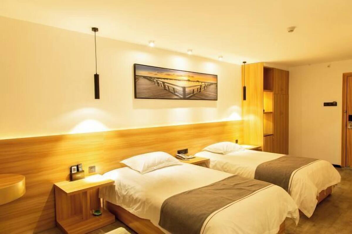 A hotel room with two beds, a wooden headboard, a painting on the wall, and a nightstand with a lamp.