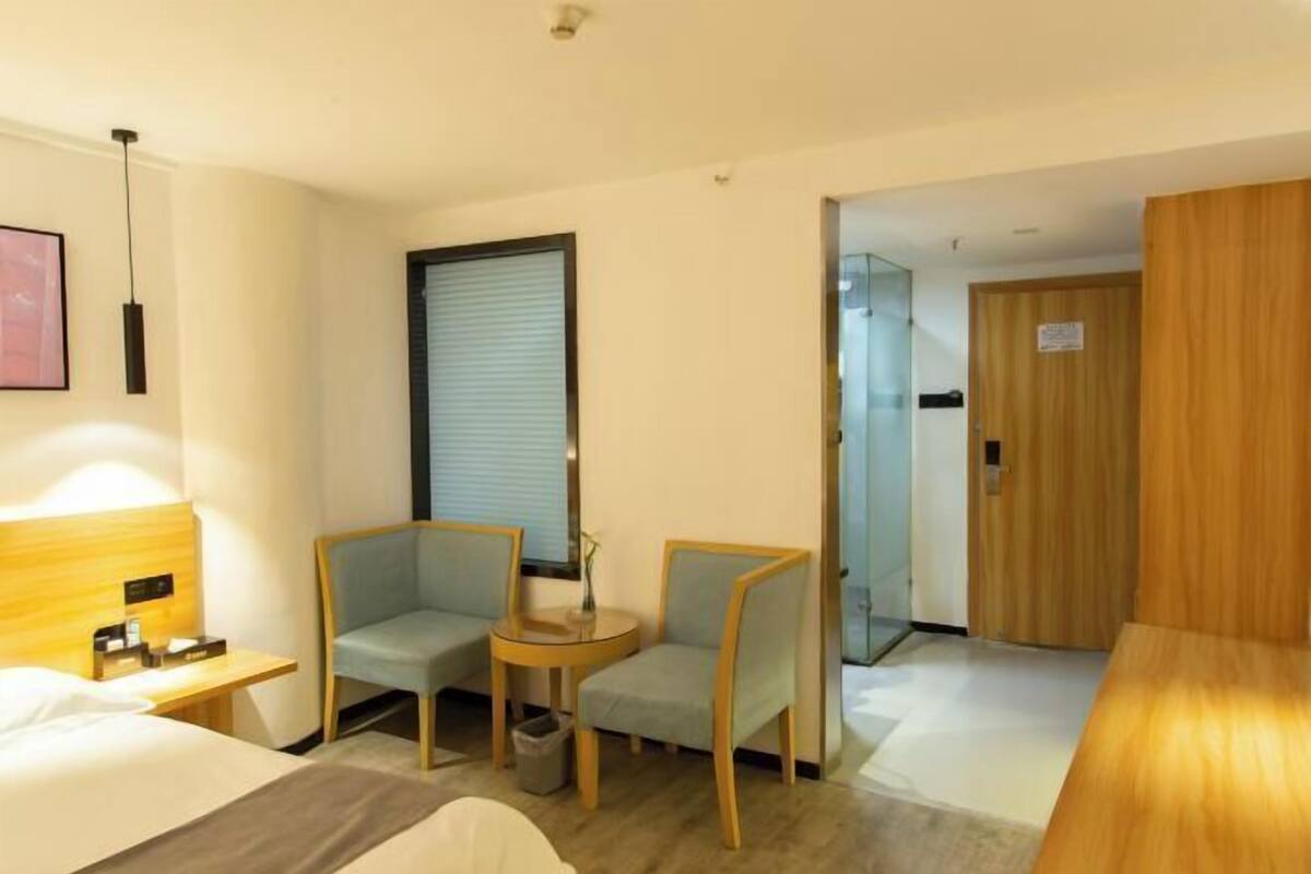 A hotel room with a bed, a small table, two chairs, a waste bin, and a door leading to another room.