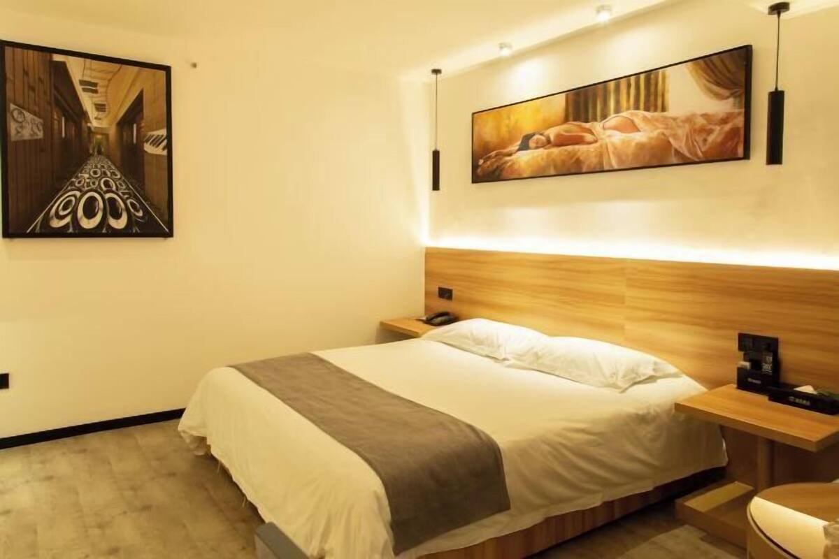 A modern hotel room with a large bed, a wooden headboard, a bedside table with a lamp, and artwork on the walls.