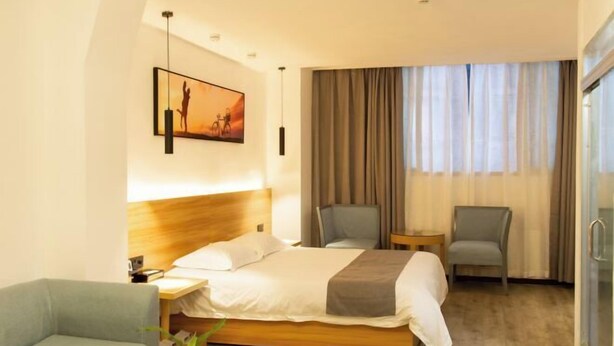 A hotel room with a bed, two chairs, a small table, a nightstand, a lamp, a painting, and a window with curtains. - Tianyi Art Hotel (Minhou)