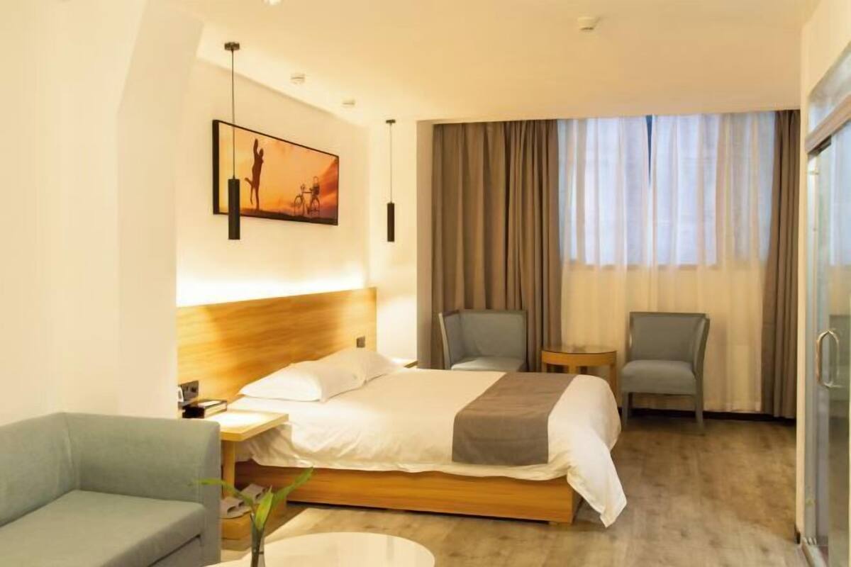 A hotel room with a bed, two chairs, a small table, a nightstand, a lamp, a painting, and a window with curtains.