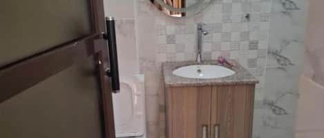 A compact bathroom with a wooden vanity, a white sink, and a mirror above it. There is a toilet to the left and a shower area to the right.