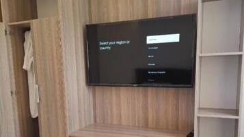 Habitación de hotel con una televisión de pantalla grande montada en un mueble de madera, cama con ropa de cama blanca y un armario con una toalla colgada.