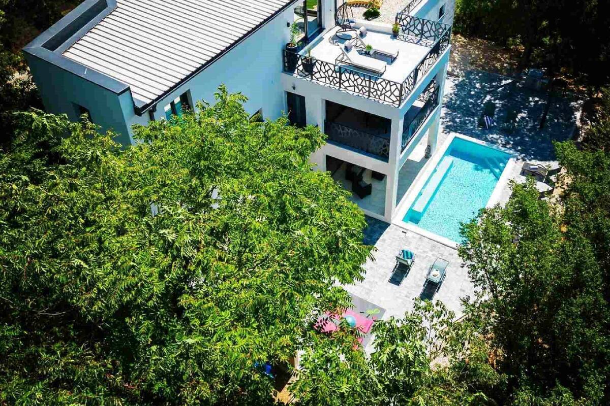Una casa moderna de dos pisos con piscina, rodeada de exuberante vegetación.