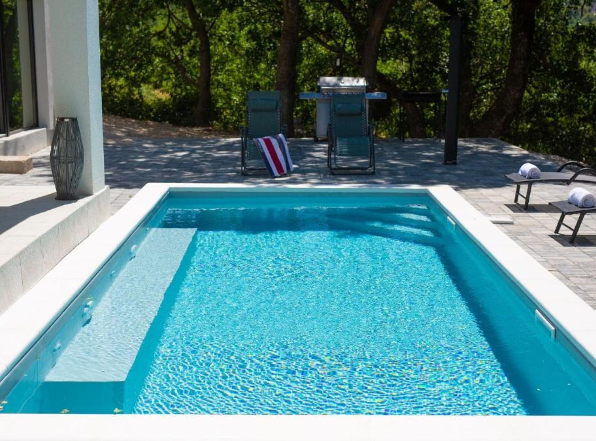 A rectangular pool with clear blue water, surrounded by a tiled deck. There are lounge chairs and a small table with a flag in the background.