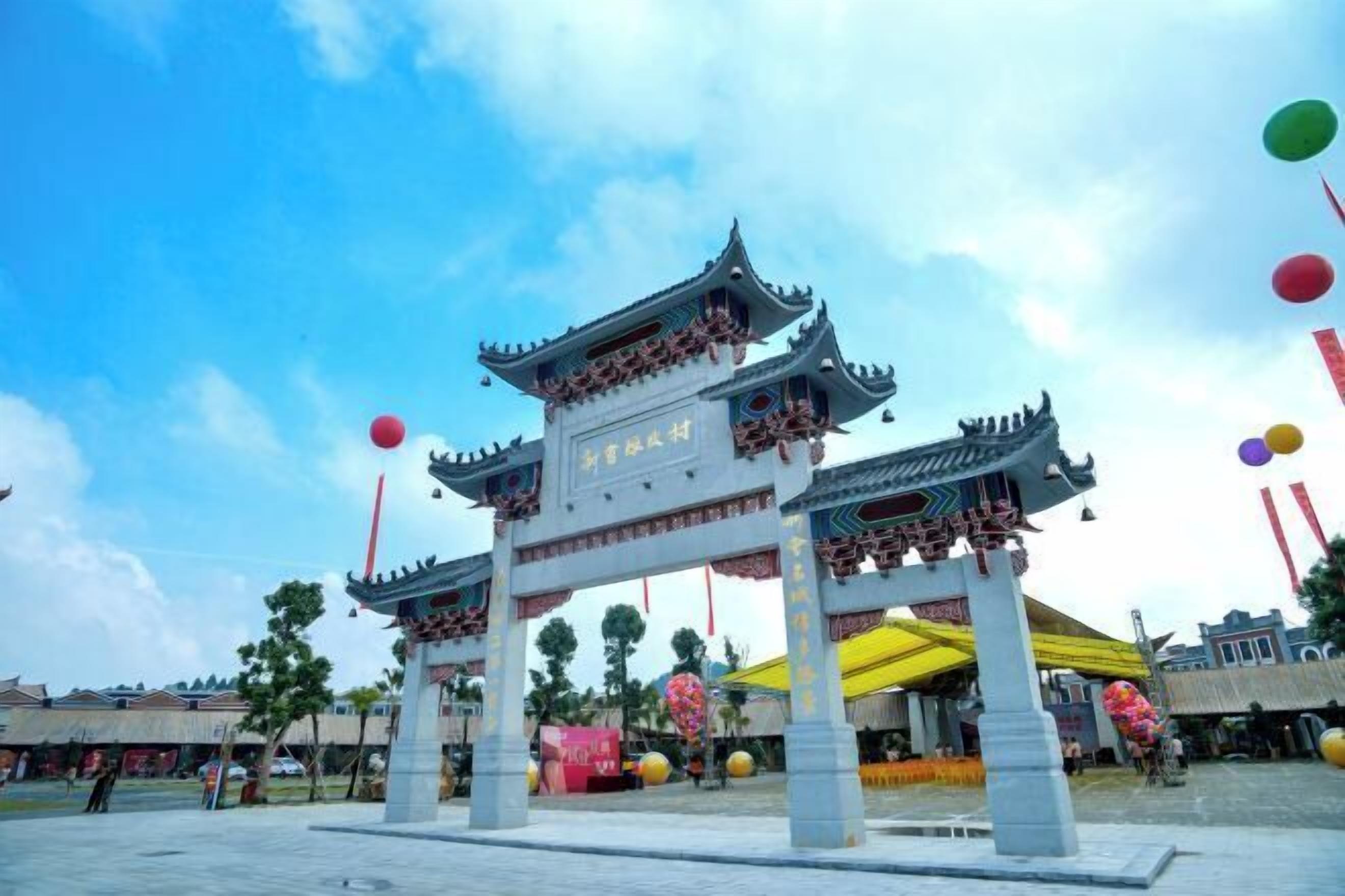 A traditional Chinese gate with intricate designs and characters, set in an open plaza with trees and a clear sky.